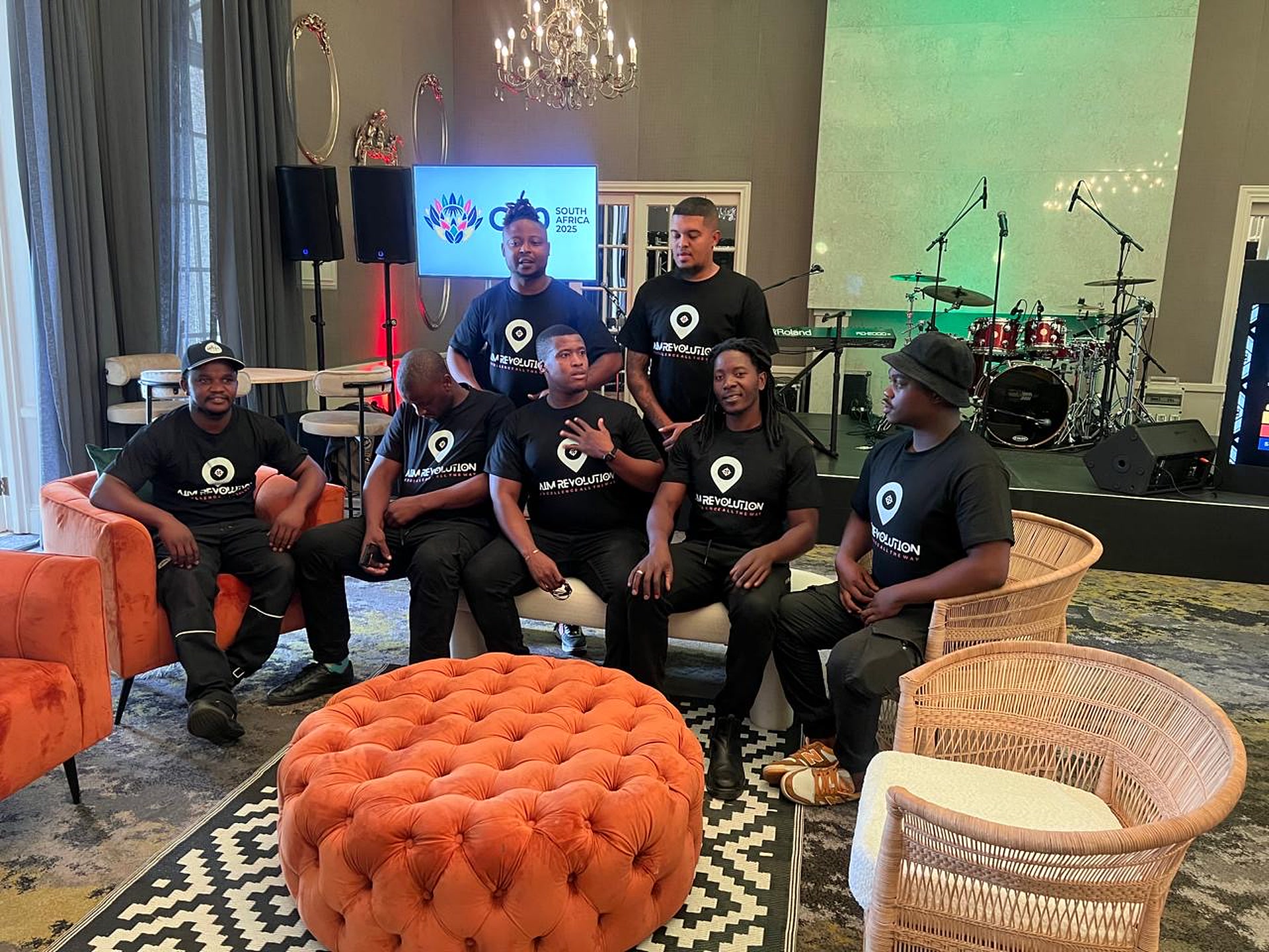 Seven members of the AIM Revolution crew in branded shirts in a lounge, with a live band stage and a G20 South Africa screen behind them